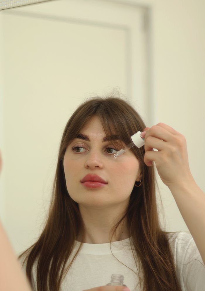 Woman applying skincare serum with a dropper in front of a mirror.