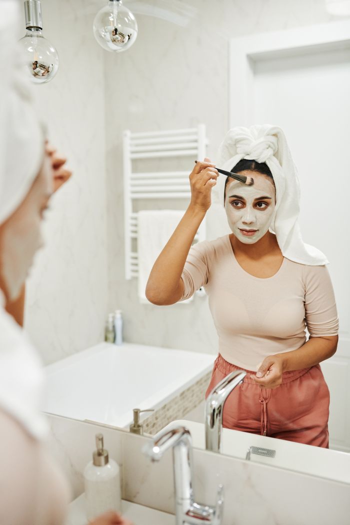 Offerings Woman applying clay mask with a brush in her bathroom mirror reflection.